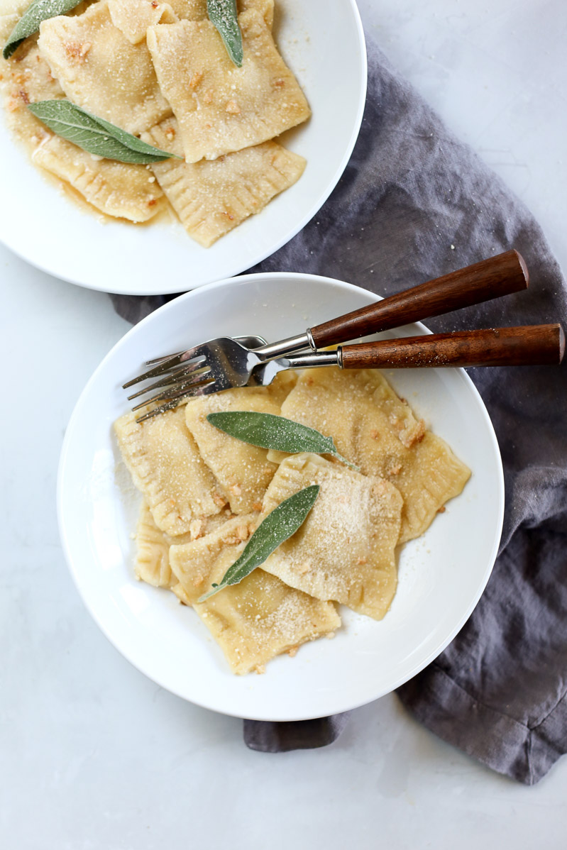 Overhead view of two plates of ravioli on a white table with a dark grey cloth. Two wooden-handle forks rest on one of the plates. Fresh sage leaves used as a garnish.