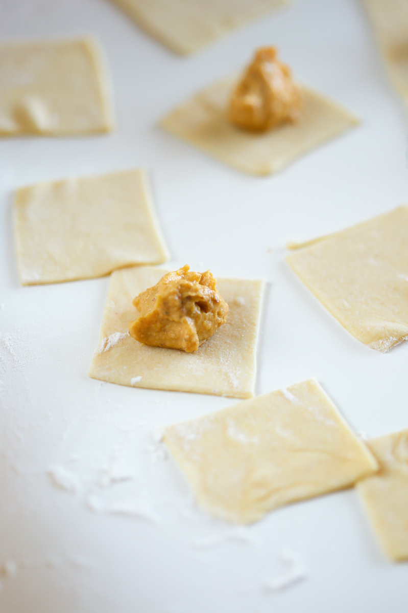 Ravioli dough cut into squares and laid on a white table, a teaspoon of butternut squash ravioli rests in the middle of a piece of dough.