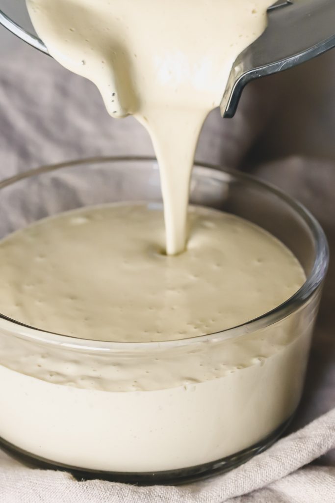 A blender pouring creamy vegan caesar dressing into a glass container.