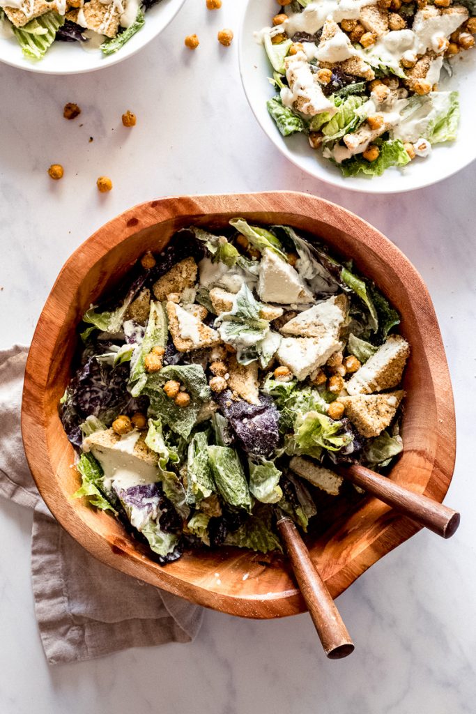 Brown wood bowl with lettuce, crispy tofu, crispy chickpeas and vegan caesar dressing drizzled on top.