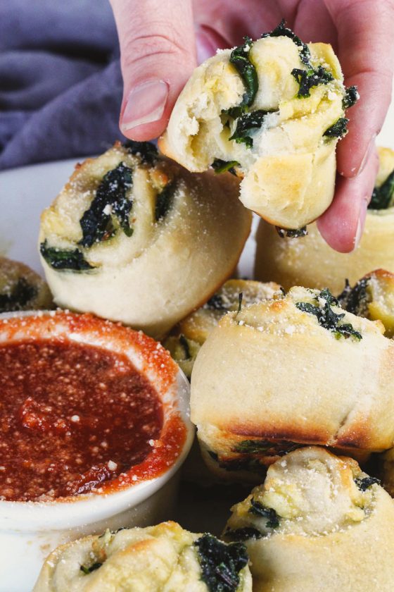 Vegan Spinach Pinwheels by Pasta-based. Close up side shot of hand holding spinach pinwheel over a plate of extra pinwheels and small white bowl of red sauce with bite taken out.