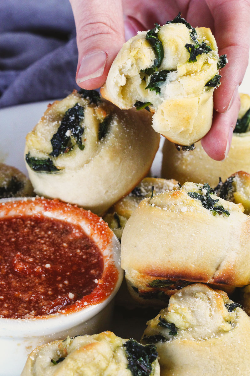 Vegan Spinach Pinwheels Recipe by Pasta-based.  Close up side shot of hand holding spinach pinwheel over a plate of extra pinwheels and small white bowl of red sauce with bite taken out.