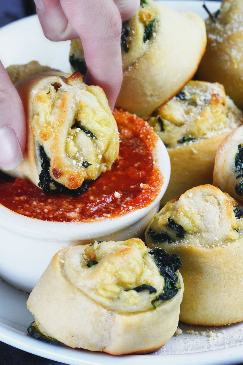 Vegan Spinach Pinwheels Recipe by Pasta-based.  Close up shot of hand dipping a vegan spinach pinwheel into a small white bowl of red sauce.  Extra pinwheels surrounding the white bowl of sauce.