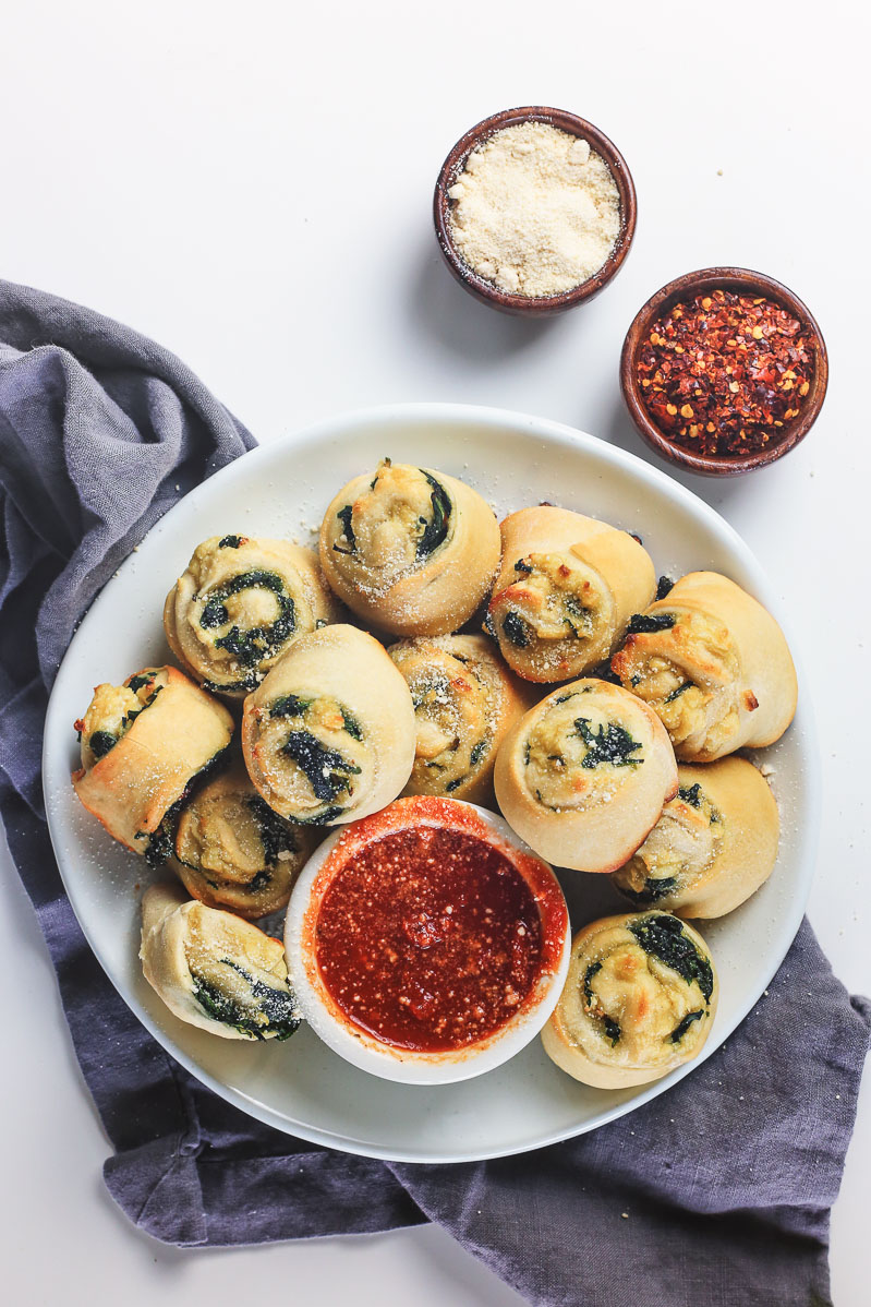 Vegan Spinach Pinwheels Recipe by Pasta-based. Overhead photo of a white plate placed on top of a gray dish towel overflowing with Vegan Spinach Pinwheels with a small white bowl of red dipping sauce.  Red pepper flakes and vegan parmesan cheese in small wooden bowls off to the top right of the plate on the white table.