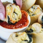 Vegan Spinach Pinwheels by Pasta-based. Close up shot of hand dipping a vegan spinach pinwheel into a small white bowl of red sauce. Extra pinwheels surrounding the white bowl of sauce.