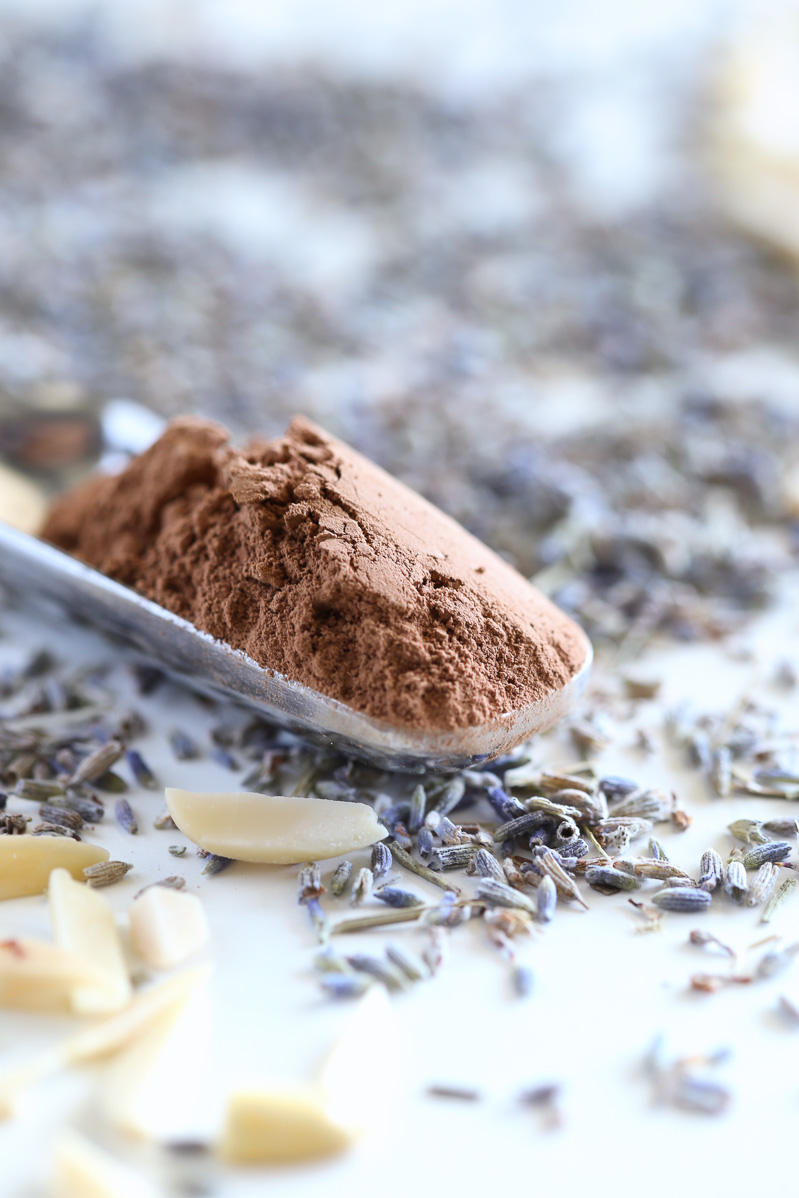 A small scoop of cocoa powder in a silver scoop, on a white table with lavender buds, and blanched almonds randomly placed around it.