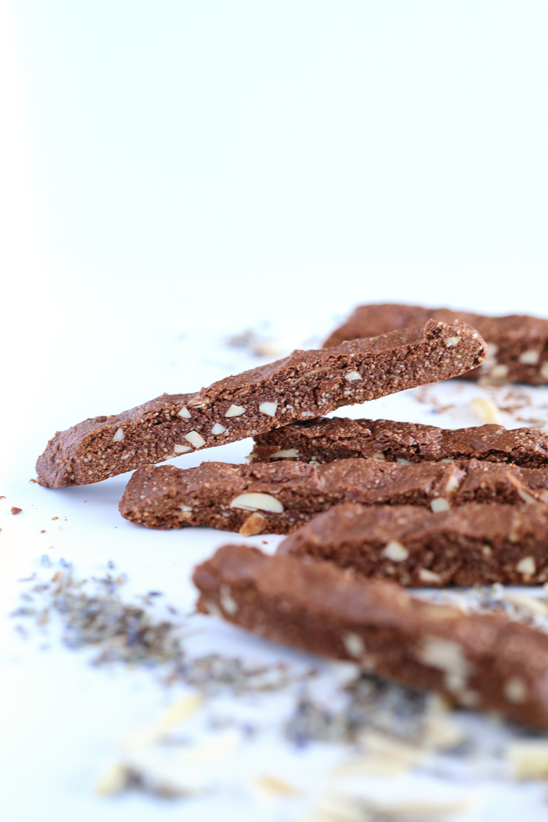 A chocolate lavender biscotti leaning on top of other biscotti on a white table. Lavender and almonds sprinkled around the table.