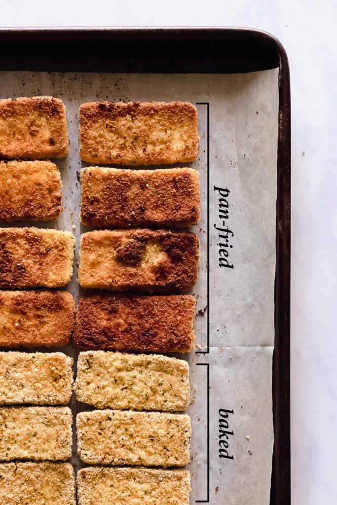 Crispy breaded vegan tofu cutlets lined up on a parchment paper lined baking sheet. Half of the breaded cutlets are deep golden brown with text next to it saying that they are pan-fried. The other half of the cutlets are baked so they are a lighter brown with text next to it saying baked.