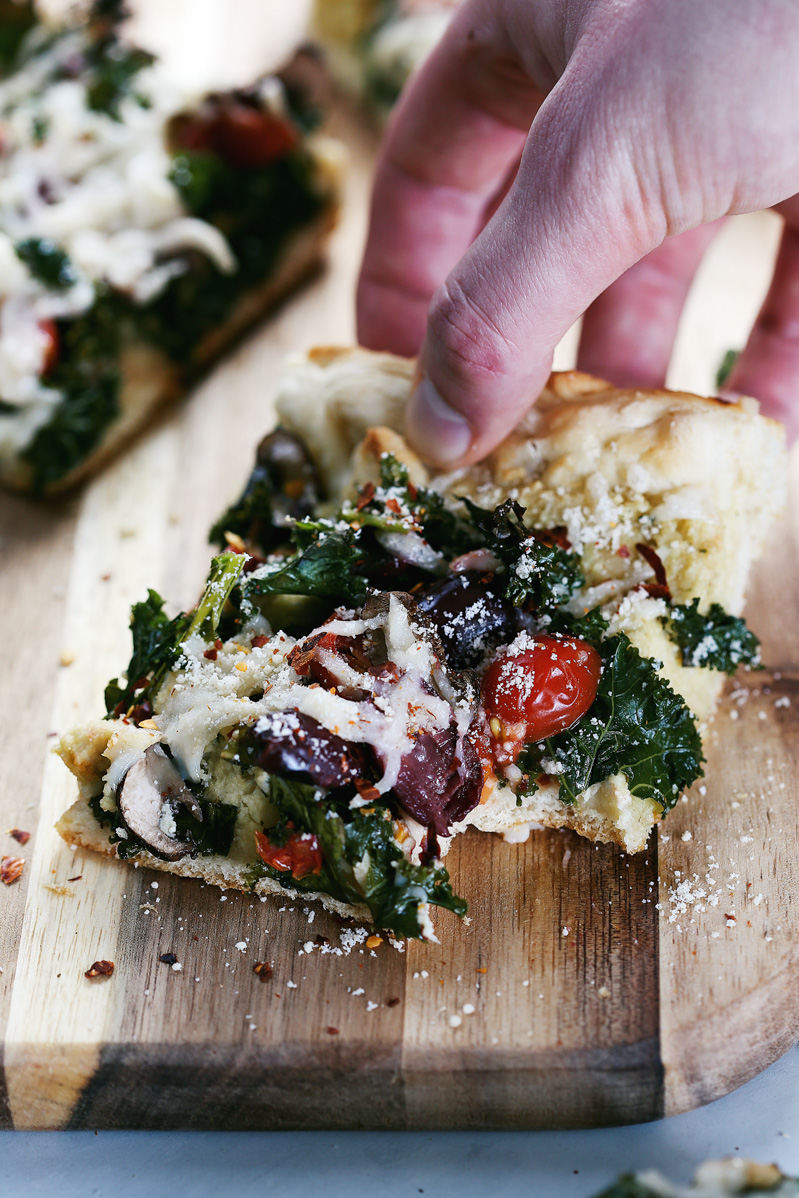 Vegan Crispy Kale Grandma-Style Pizza by Pasta-based. A hand reaches in to put down a slice of pizza with a bite taken out of it.