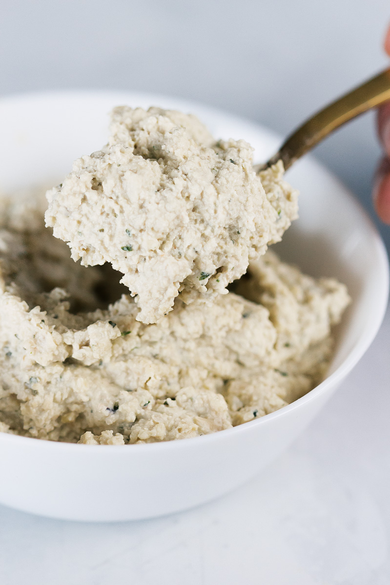 Vegan Crispy Kale Grandma-Style Pizza by Pasta-based. A photo of the cashew ricotta cheese. A spoon with a scoop of cheese hovers over the bowl of ricotta.