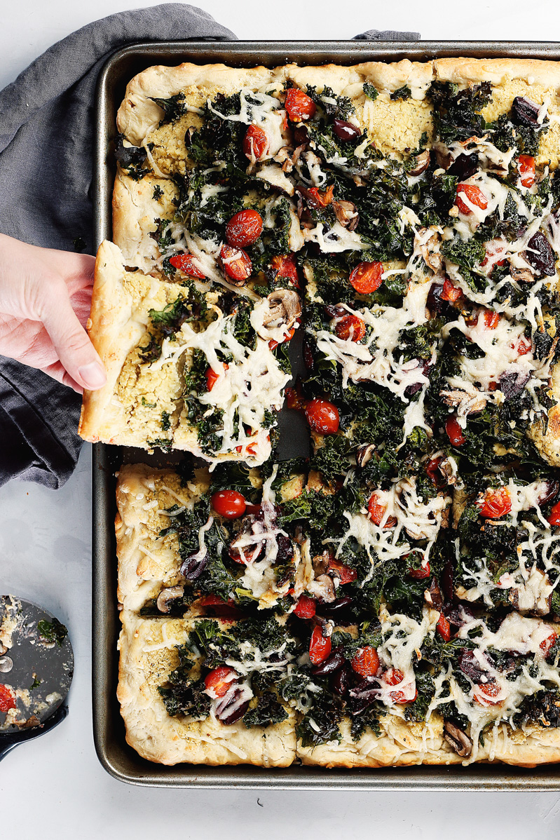 Vegan Crispy Kale Grandma-Style Pizza by Pasta-based. Made in a cookie sheet, this rectangular pizza is topped with crispy kale, homemade cashew ricotta, vegan mozzarella, grape tomatoes, mushrooms, and kalamata olives.