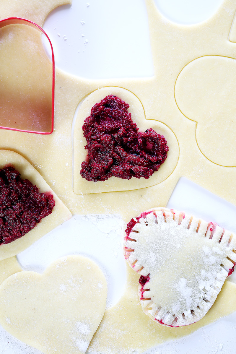 Vegan Red Beet Pesto Ravioli by Pasta-based. The making of red beet pesto ravioli, heart-shapes cut into vegan ravioli dough and a spread of pesto in the middle of each cut of dough. One ravioli already sealed with crimps of fork in the corner of the photo.