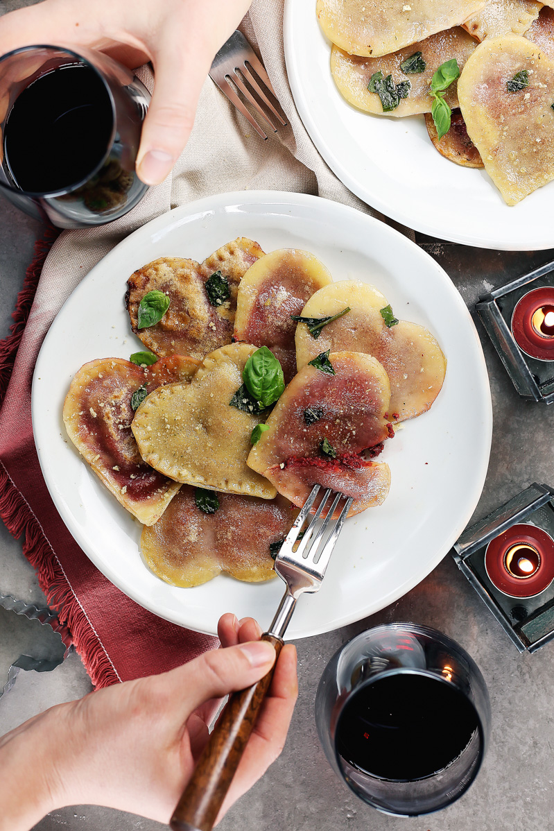 Vegan Red Beet Pesto Ravioli by Pasta-based. A dinner table set with two plates of heart-shaped red beet pesto ravioli, tea-light candles, and red wine.