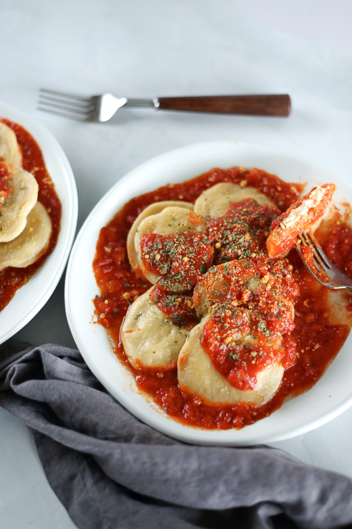 A white plate with ravioli covered in red sauce and parsley garnish.  One ravioli is cut in half to show vegan ricotta cheese inside.