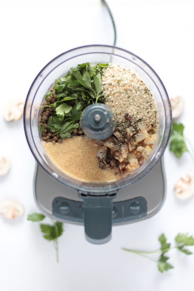 Overhead view of the food processor with the meatball ingredients in it, not yet mixed. Breadcrumbs, mushrooms, onions, lentils, parsley and a flax egg. Garlic, parsley and mushrooms are scattered around in the blurry background.