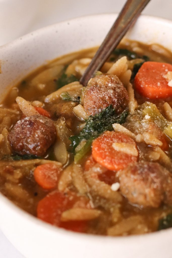 closeup of vegan meatballs inside white bowl of soup with a spoon sticking out.  Carrots and orzo floating around with escarole there as well.