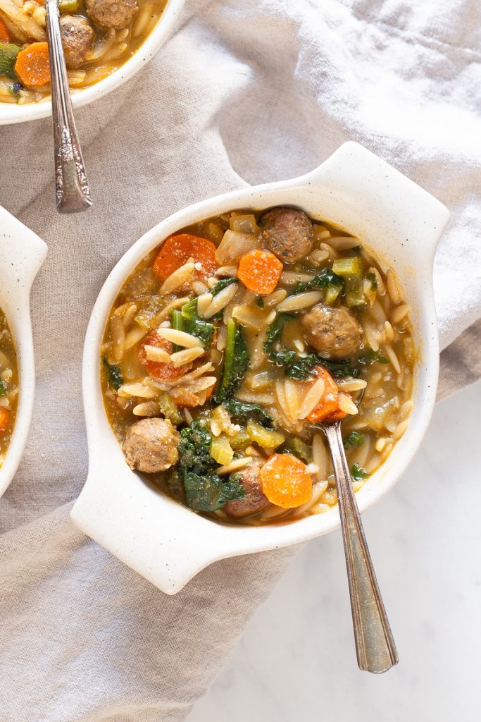 overhead photo of vegan meatballs inside white bowl of soup with a spoon sticking out.  Carrots and orzo floating around with escarole there as well.