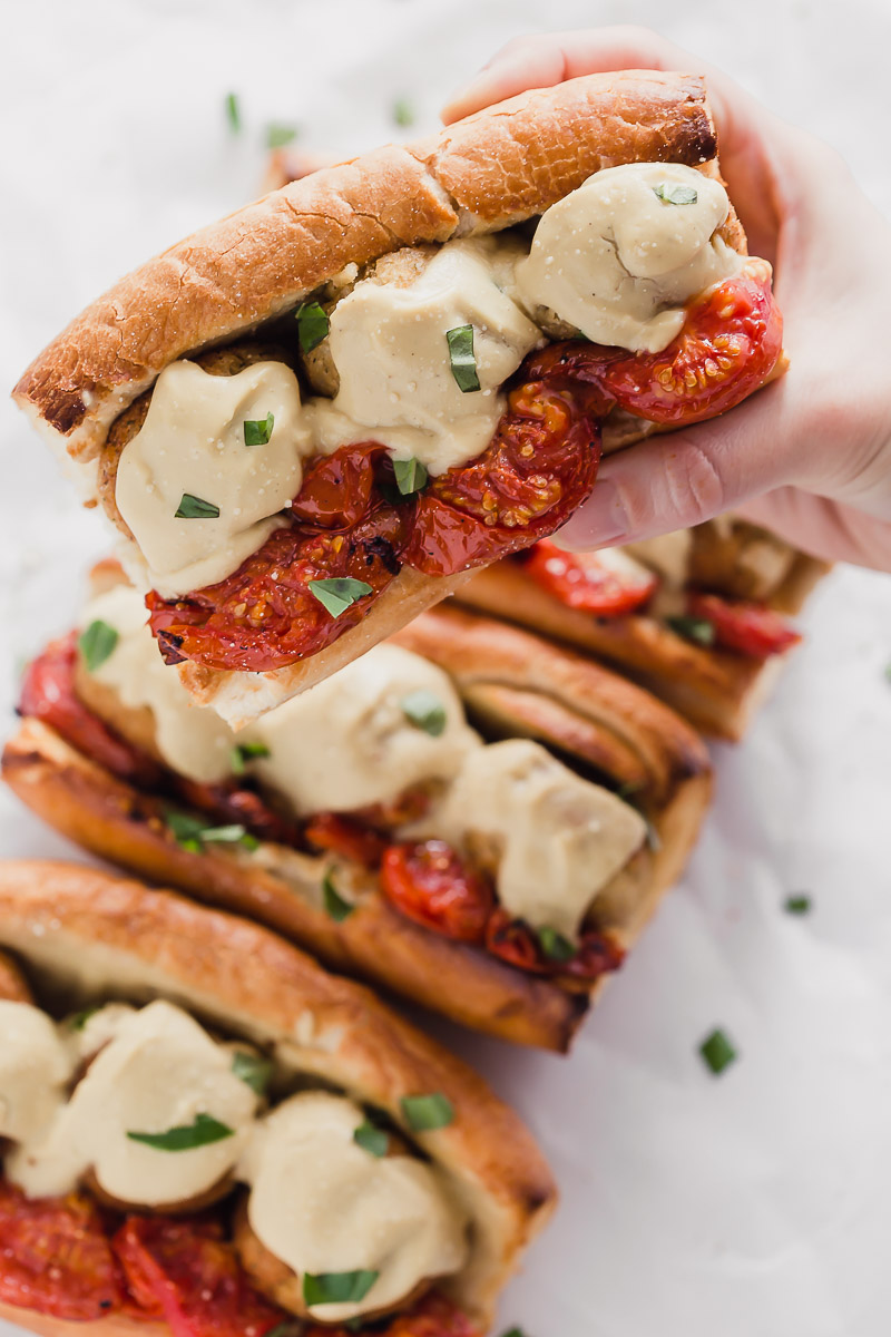 Vegan Meatball Subs by Pasta-based. Someone holding up a juicy meatball hero on toasted bread with roasted campari tomatoes, vegan cauliflower alfredo sauce, and cauliflower brown rice meatballs. Fresh basil sprinkled on top.