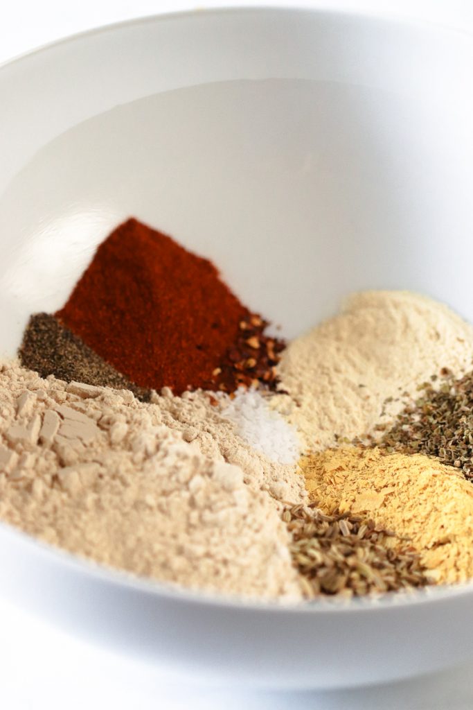 A white mixing bowl with all the dry ingredients piled up along side each other.  Vital Wheat Gluten, paprika, garlic powder, nutritional yeast, fennel, oregano, salt and pepper.