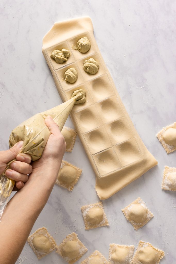 A sheet of ravioli dough on top of a ravioli mold tray.  Hands are holding a pastry bag full of vegan ricotta and filling in the ravioli divots.