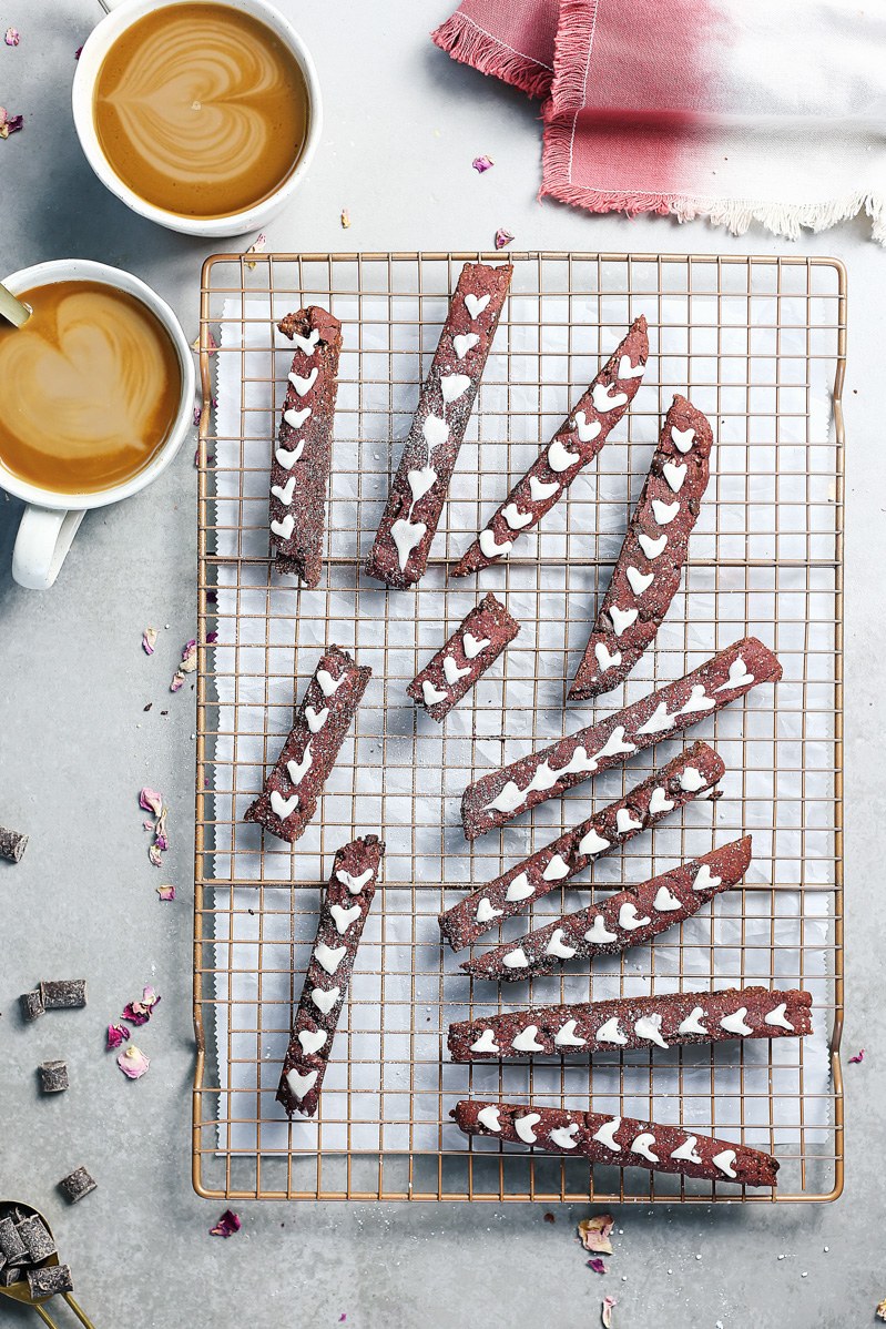 Red Velvet Biscotti by Pasta-based. A wide overhead shot of the biscotti laying on a cooling rack with white icing hearts. Two coffees are on the table waiting to be dipped into.