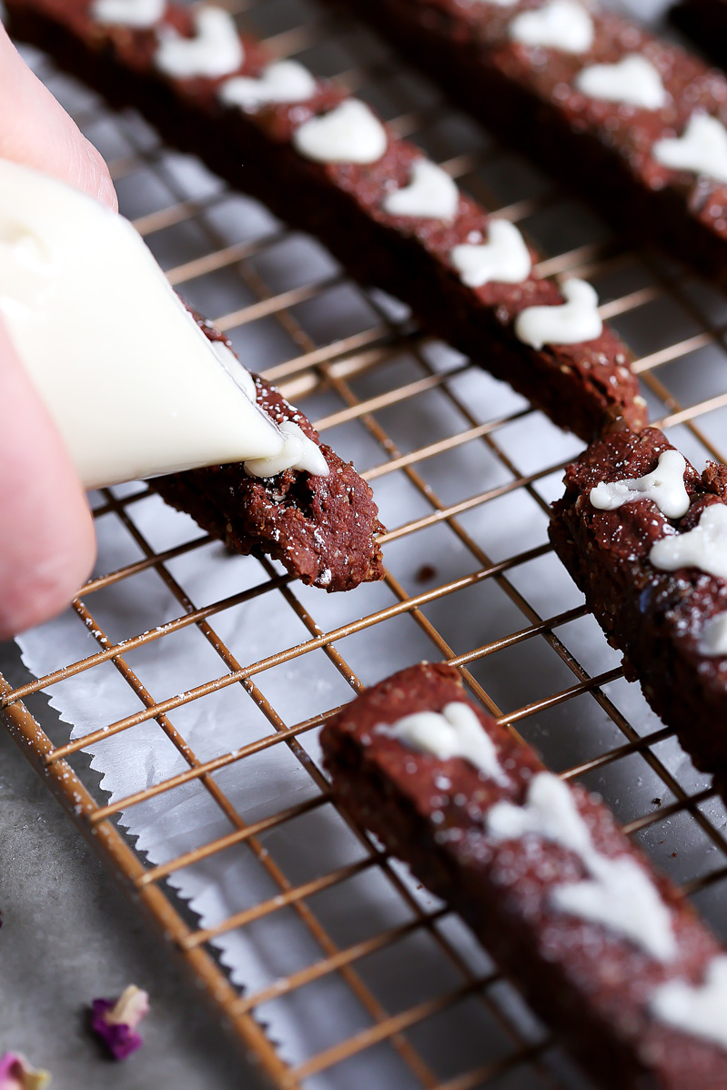 Red Velvet Biscotti by Pasta-based. Piping hearts with icing onto the cookies. The icing is simply made from powdered sugar and plant milk.