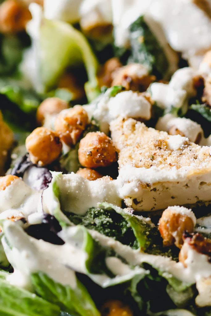 Close up of lettuce, crispy tofu, crispy chickpeas and vegan caesar dressing drizzled on top.