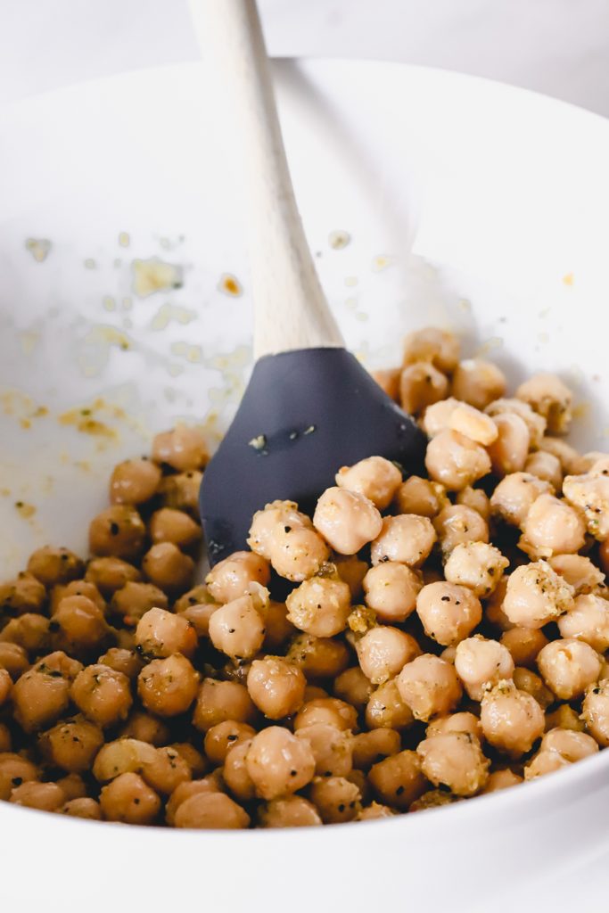 A mixing bowl of chickpeas with oil/herbs coating them and a silicone spatula.