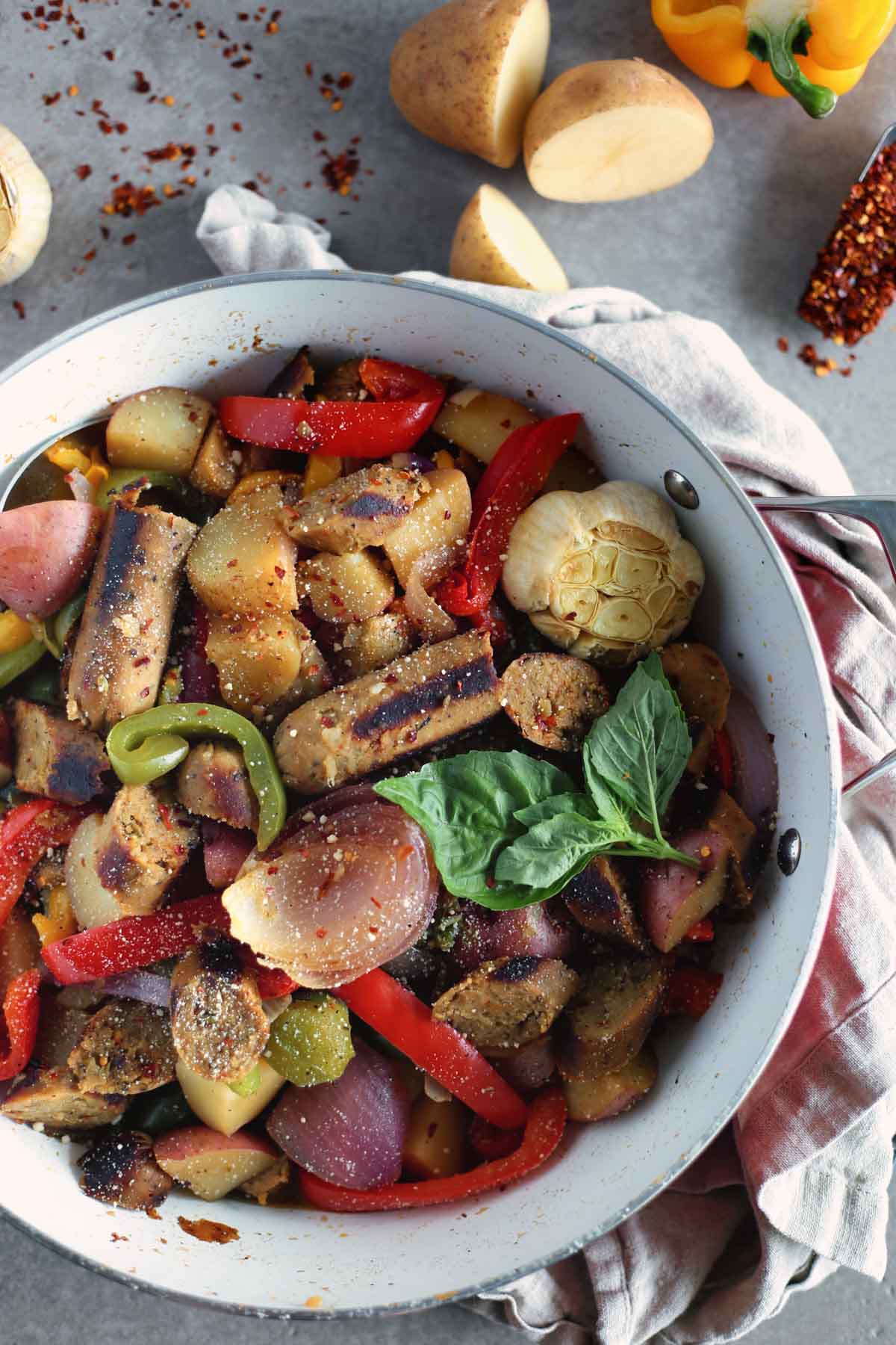 Vegan Sausage, peppers, onions and potatoes in a white pan overhead with Potatoes, chopped pepper and roasted garlic in background.