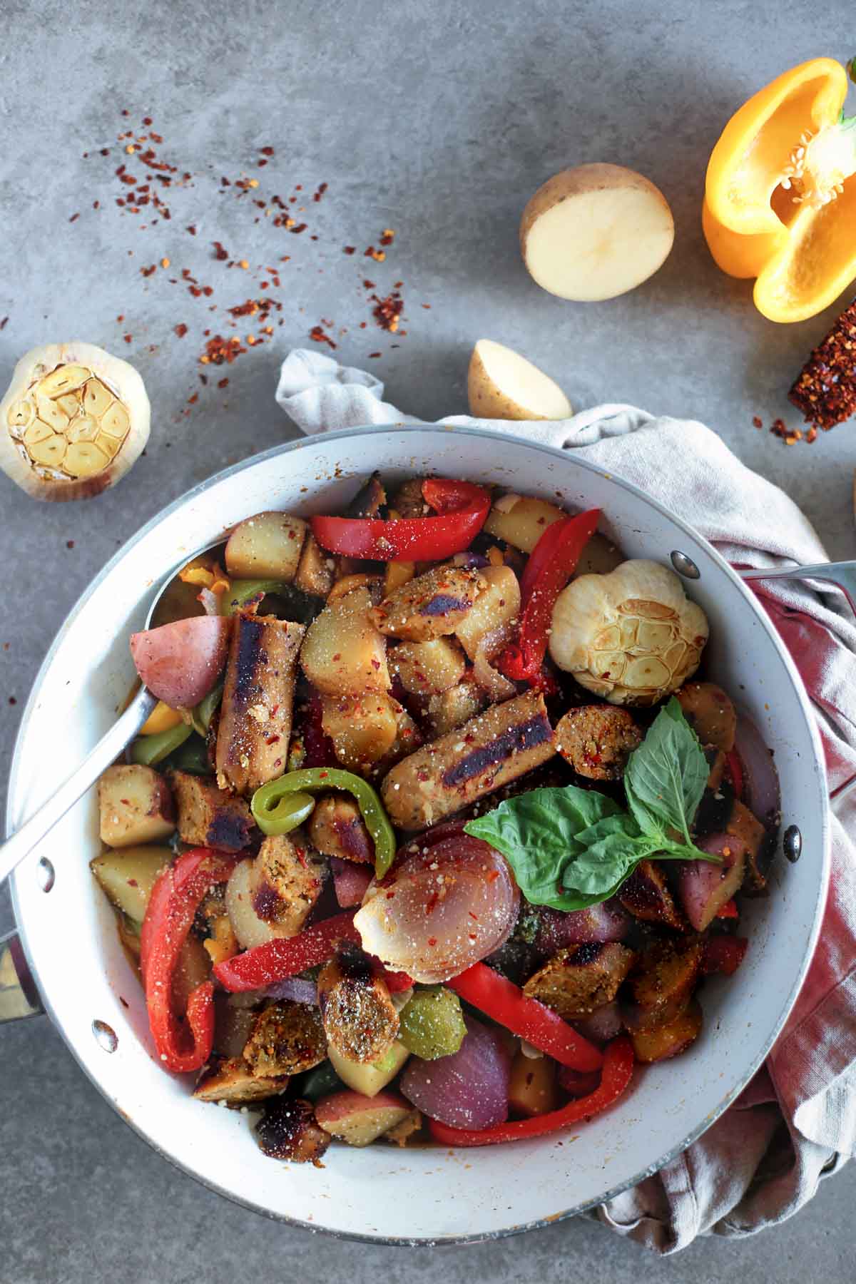 Vegan Sausage, peppers, onions and potatoes in a white pan overhead with Potatoes, chopped pepper and roasted garlic in background.