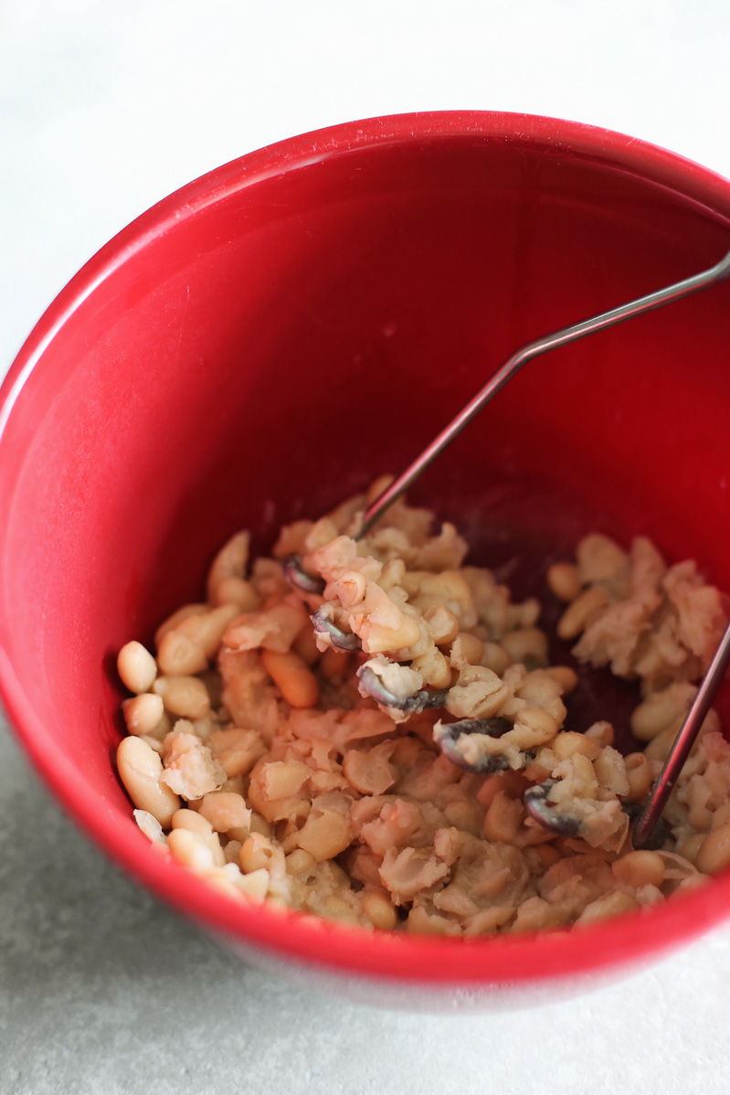 Vegan Seitan Parmesan by Pasta-based. A red bowl with cannellini beans inside. A potato masher leaning inside the bowl with some of the beans already mashed up.