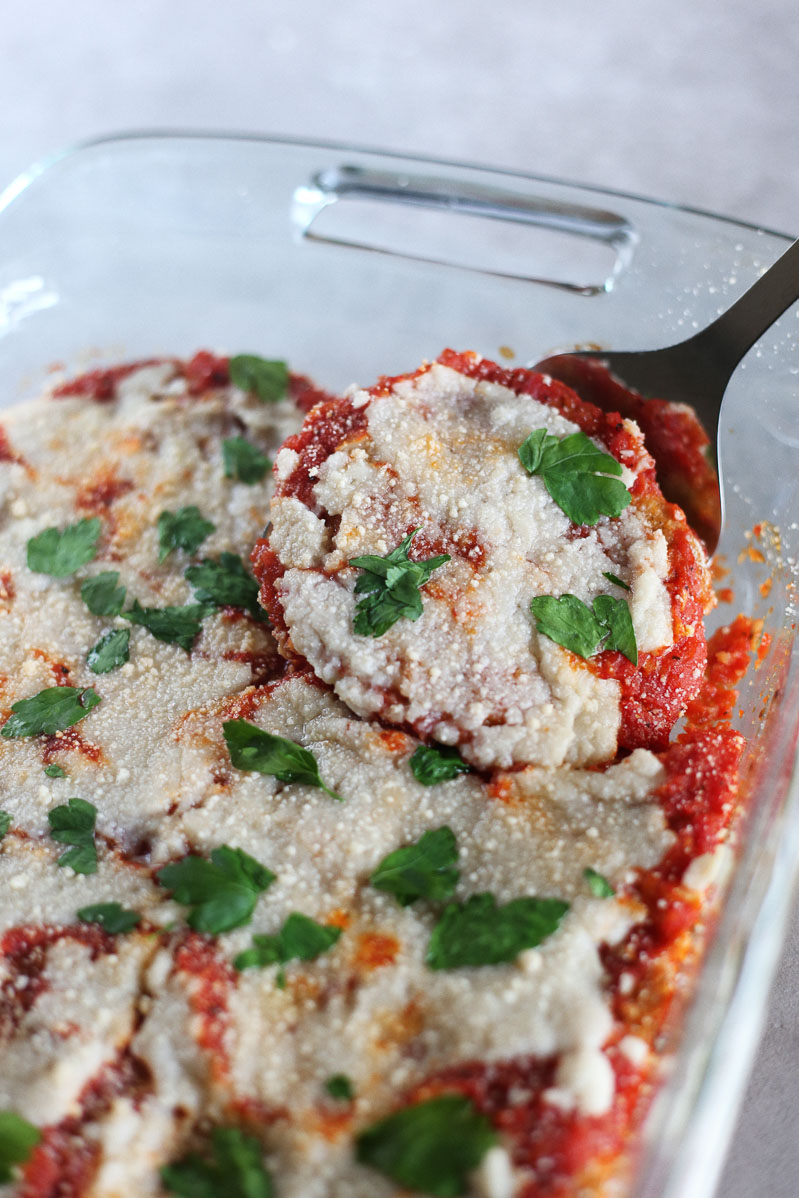 Vegan Seitan Parmesan by Pasta-based. Seitan cutlets smothered with marinara sauce and topped with melted vegan mozzarella cheese in a baking dish with a spatula lifting one piece out of the pan.