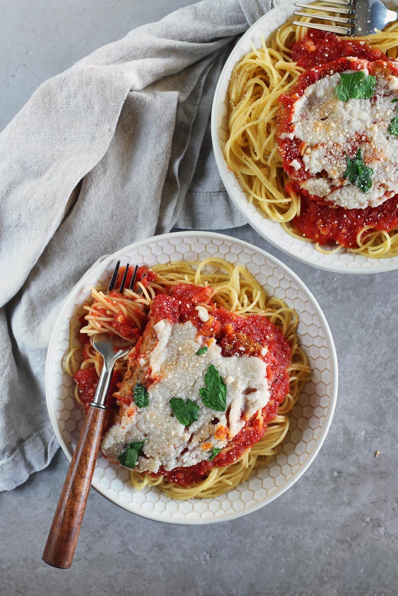 Vegan Seitan Parmesan by Pasta-based. An overhead shot of two bowls of vegan seitan parmesan smothered in marinara sauce and topped with vegan melted mozzarella cheese. On a grey countertop with a light grey napkin. A fork with a wooden handle resting in one of the bowls.