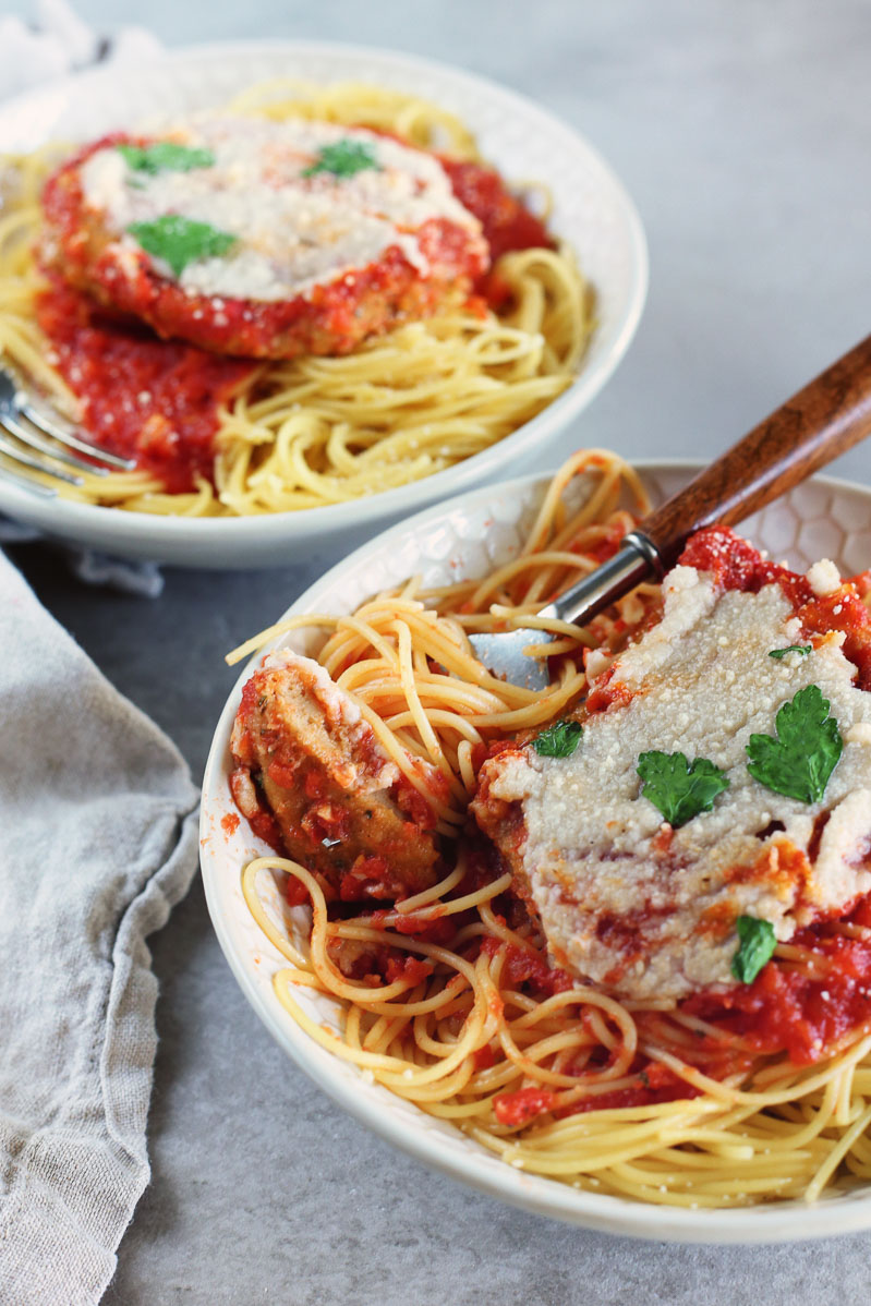 Vegan Seitan Parmesan by Pasta-based. Bowls of spaghetti with seitan cutlets smothered in marinara sauce and topped with vegan mozzarella cheese. A fork with a piece of seitan leaning on the side of the bowl.