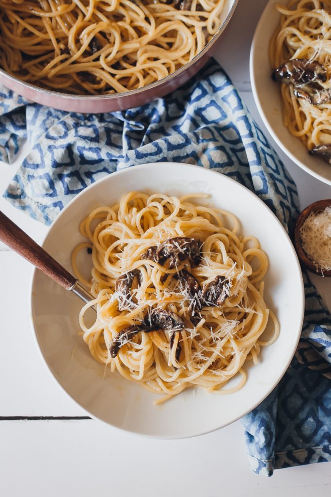 A white bowl filled with Vegan Spaghetti Carbonara with Mushroom Bacon. A blue cloth with a diamond pattern is underneath the bowl. A fork with a wooden handle is placed inside the bowl from the left side. At the top, there is a pan with some extra Vegan Spaghetti Carbonara.