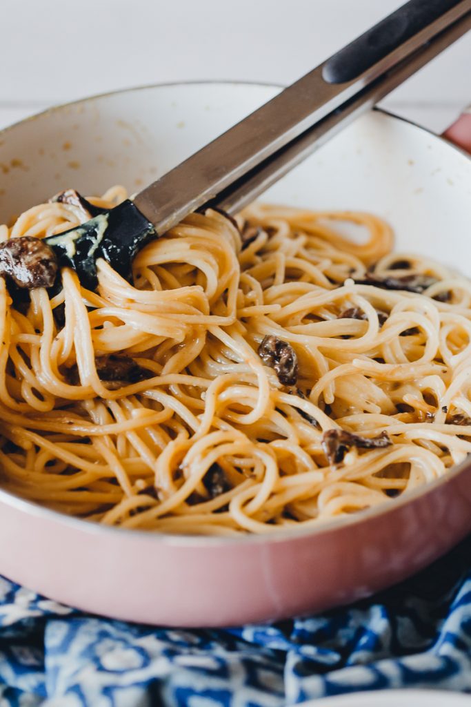 Vegan Spaghetti Carbonara by Pasta-based. Creamy spaghetti noodles in a pink saute pan with a blue cloth underneath. Stainless steel tongs are resting in the pan with spaghetti tangled between them. Marinated mushrooms are peeking through the spaghetti.