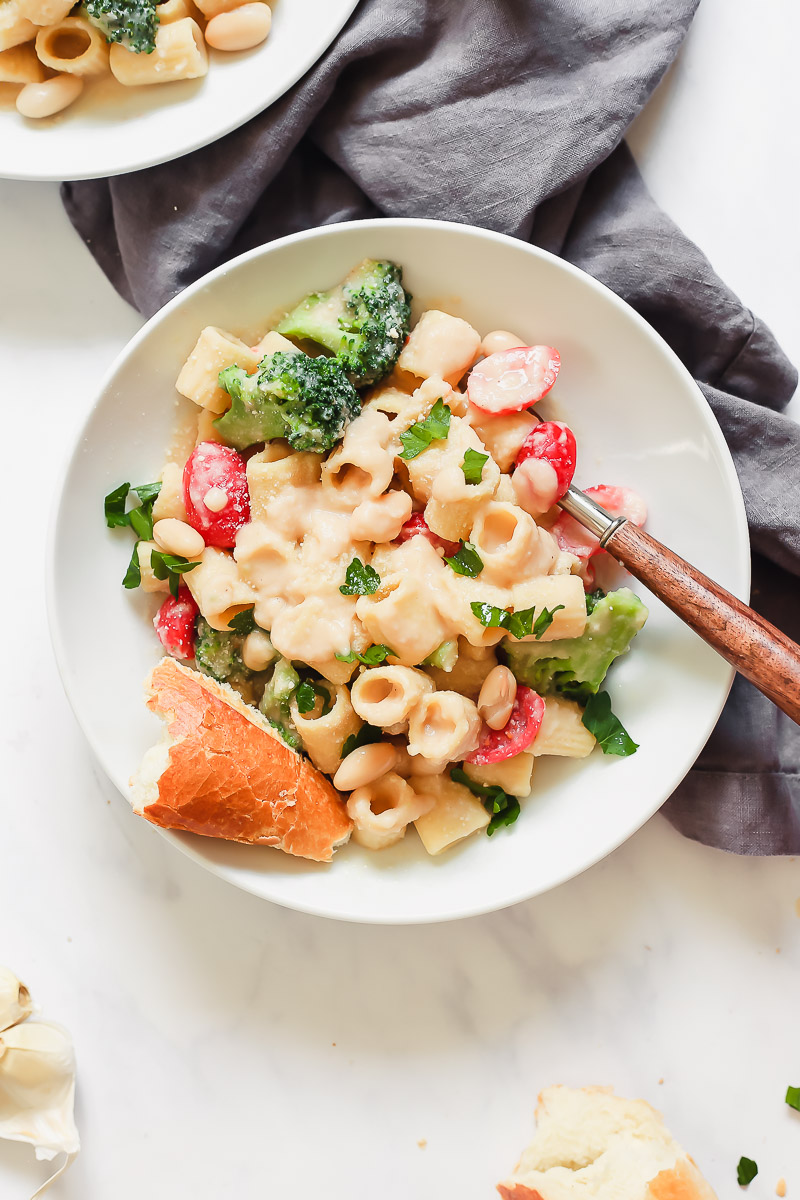 Vegan White Bean Alfredo by Pasta-based. Creamy, cheesy, healthy, low-calorie and low-fat vegan alfredo sauce made with white beans, garlic, and vegetable broth. Two white bowls filled with mezze rigatoni pasta, smothered in creamy white bean alfredo sauce and tossed with fresh broccoli and grape tomatoes.